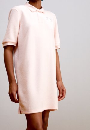 Osoba v lehkých růžových polokošilových šatech s krátkými rukávy stojící před jednobarevným šedým pozadím, ruce volně podél těla.