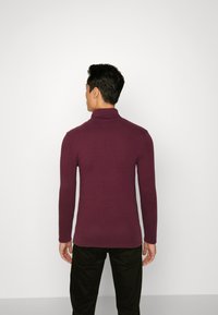 Suéter de cuello alto burdeos de manga larga, hecho de una tela suave y elástica. Presenta un diseño ajustado con hombros sin costuras y detalles de costura mínimos.