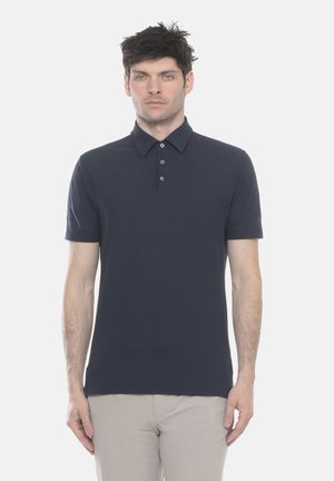 Uomo in piedi di fronte, indossa una polo blu navy a maniche corte e pantaloni beige chiaro su uno sfondo bianco uniforme.