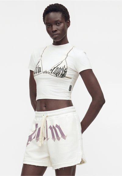 Γυναίκα φοράει λευκό cropped T-shirt με μαύρο τρίγωνο σχέδιο και λευκά σορτς με κορδόνι και μεγάλο μωβ κείμενο, στέκεται σε απλό φόντο.