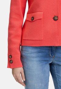 Veste en tricot corail avec deux poches avant, boutons marron et coupe ajustée. Associée à un jean en denim bleu clair.