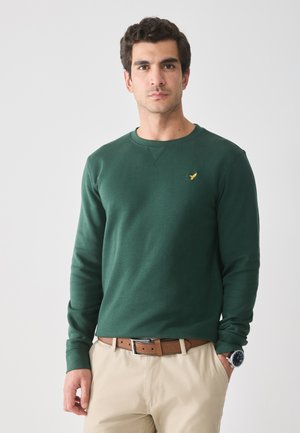 Man in groene sweatshirt en beige broek staat met één hand in zijn zak tegen een eenvoudige witte achtergrond.