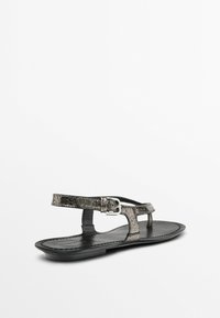 Ploché černé sandály s metalickým popraskaným vzorem, designem T-strap a nastavitelnou přezkou. Hladká kožená stélka a texturovaná podrážka.