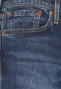 Gros plan sur un jean en denim bleu montrant la poche avant, la petite poche à monnaie avec rivets en cuivre et les détails de couture dorée.