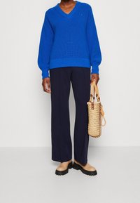 Pull en maille bleue à encolure en V, associé à un pantalon large bleu marine. Accessoire : sac en paille tressée avec des accents en cuir camel et des poignées.