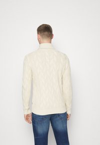 Cremefarbener Kabelstrickpullover mit hohem Kragen, strukturiertem Muster sowie gerippten Bündchen und Saum, kombiniert mit blauen Jeans.