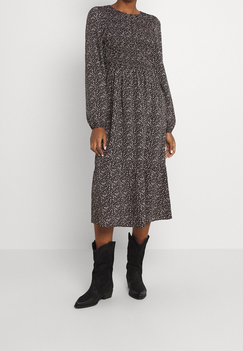 Schwarzes florales Midikleid mit langen Ärmeln, strukturiertem Stoff und gerafftem Taillenbund. Kombiniert mit schwarzen Cowboy-Stiefeln.