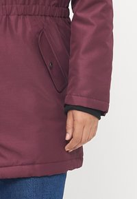 Casaco isolado cor bordeaux com cintura ajustada, um bolso lateral e um punho preto. O tecido parece suave e resistente à água.