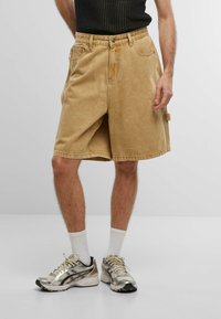 Shorts de mezclilla color beige con un ajuste relajado, que cuentan con bolsillos frontales y una tira lateral. Llevados con calcetines blancos y zapatillas de running metálicas.