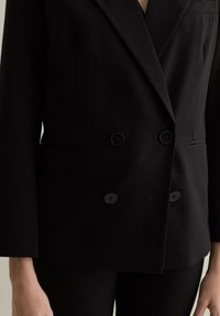 Blazer noir à double boutonnage en tissu lisse, avec deux boutons, des poches latérales et un design de col classique.