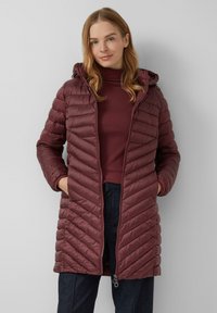 Burgundská péřová bunda s prošívaným vzorem šípů, kapucí, předním zipem a bočními kapsami, oblečená přes ladící top a tmavé kalhoty.