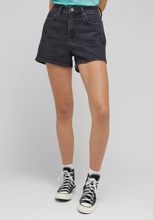 Personne portant un short en jean noir, des baskets montantes noires avec des lacets blancs et des chaussettes blanches aux chevilles, se tenant devant un fond uni.