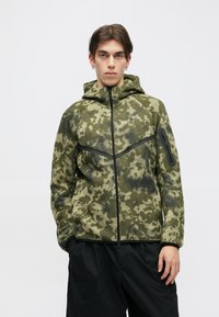 Zaļš kamuflāžas jaka ar rāvējslēdzēju un kapuci, izgatavota no tekstūru auduma, ar melniem akcentiem un rāvējslēdzēju kabatu uz kreisā roka.