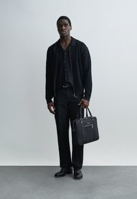 Homme portant une chemise noire, un pull à fermeture éclair noir, un pantalon noir, des chaussures noires, tenant une mallette noire, debout devant un fond gris clair uni.
