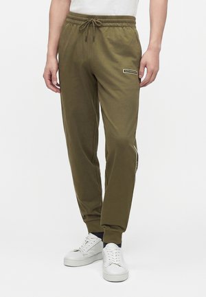 Mand iført olivengrønne joggers med snøre i taljen og hvide sneakers, stående mod en enkel hvid baggrund.