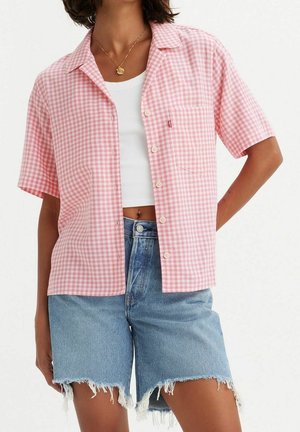 Personne portant une chemise à manches courtes à carreaux rose et blanc par-dessus un crop top blanc et un short en jean aux bords effilochés.