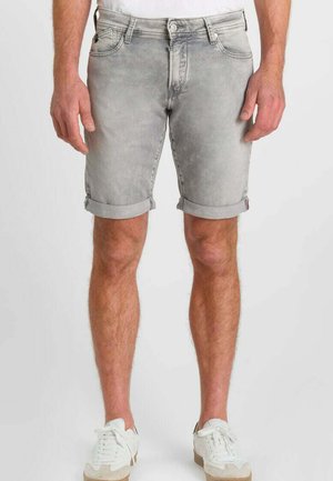 Uomo con pantaloncini di jeans grigio chiaro con orlo arrotolato e sneakers bianche, in piedi davanti a uno sfondo uniforme.