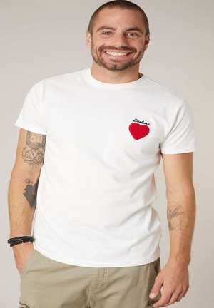 Homme souriant portant un t-shirt blanc avec un cœur rouge et le texte "Deeluxe" sur la poitrine, un pantalon beige et des tatouages visibles sur les deux bras.