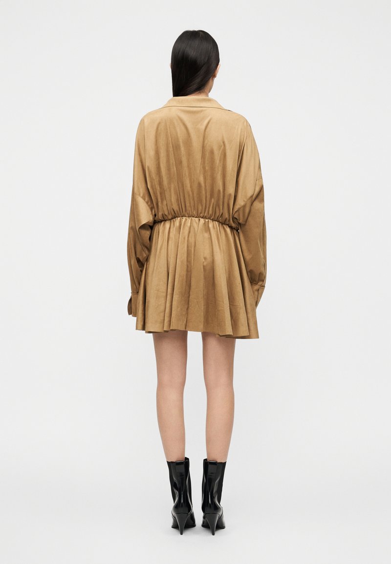 Beiges T-Shirt-Kleid mit langen Ärmeln, geraffter Taille, fließendem Rock und glatter Textur. Kombiniert mit glänzenden schwarzen Stiefeletten.