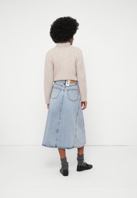 Kvinde med krøllet hår iført en beige turtleneck trøje, lyseblå denim midi-nederdel, grå sokker og sorte sneakers, stående med ryggen til.