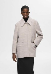 Lys grå tekstureret uldfrakke med bred krave, frontlukning med knapper og to sidelommer, båret med en sort turtleneck under.