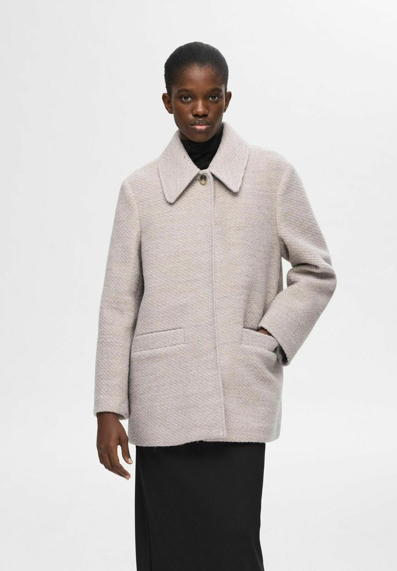 Lys grå tekstureret uldfrakke med bred krave, frontlukning med knapper og to sidelommer, båret med en sort turtleneck under.