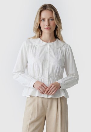 Blondynka ubrana w białą, zapinaną na guziki bluzkę z kwiatowym haftem oraz beżowe spodnie, stojąca na jednolitym jasnym tle.