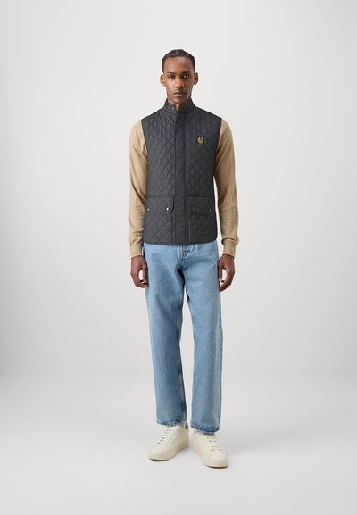 Gilet matelassé gris foncé avec col haut, doté de deux poches avant et d'un emblème de logo, porté sur une chemise beige à manches longues et un jean bleu.