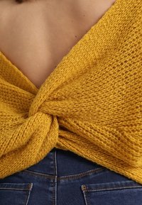 Knit yellow wrap top with a textured pattern, featuring a knotted detail at the back, worn over blue jeans with pockets visible.