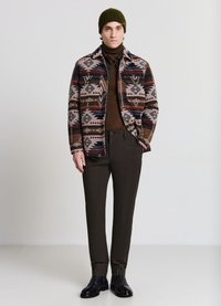 Gemusterte Herrenjacke in Braun-, Creme- und Blautönen; brauner Rollkragenpullover; dunkelbraune Hose; schwarze Schuhe; grüner Strickmütze.