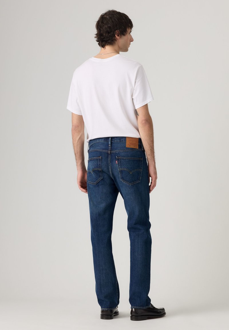 Man facing away wearing a plain white t-shirt, dark blue jeans with rear pockets, and black shoes on a plain light background.