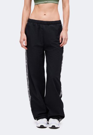 Kvinde iført sorte vidde bukser med mønstrede sidelister, hvide sneakers og en grønstribet crop top, stående mod en ensfarvet baggrund.