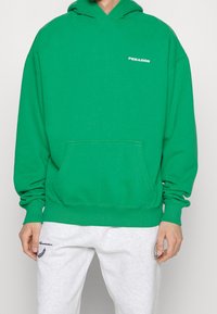 Felpa verde con tasca frontale e polsini a costine. Presenta il logo "PEGAADOR" in bianco sul petto. Indossata con pantaloni di tuta grigi.
