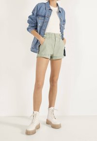 Chaqueta vaquera sobre un suéter blanco, combinada con pantalones cortos verde claro con bolsillos laterales. Botines blancos con cordones y suelas gruesas.