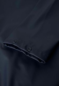 Marinblå vanntett stoff med en glatt tekstur, som har to svarte trykknapper ved mansjetten for lukking.