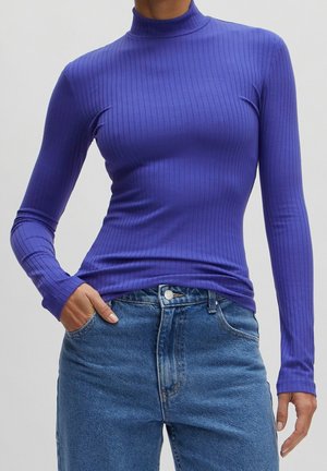 Personne portant un haut violet à col roulé côtelé, ajusté à manches longues, rentré dans un jean bleu en denim taille haute, avec une main dans la poche.