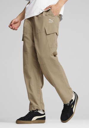Pantaloni cargo beige con tasche laterali, realizzati in tessuto leggero, abbinati a sneakers nere con accenti crema e una suola in gomma testurizzata.