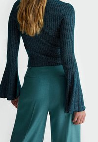 Strickoberteil in Teal mit ausgestellten Ärmeln und gerippter Textur, kombiniert mit passenden weit geschnittenen Hosen mit seitlichen Taschen.