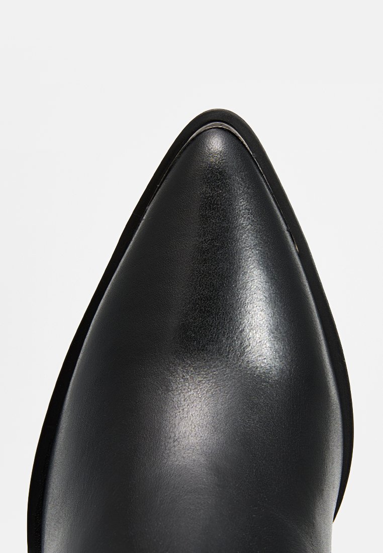 Chaussure noire en cuir à bout pointu avec une texture lisse et des coutures nettes le long du bord, posée sur un fond blanc uni.