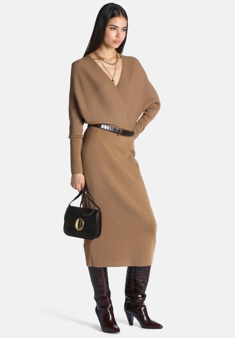 Femme portant un haut cache-cœur côtelé marron et une jupe midi, tenant un sac à main noir, accessoirisée avec des colliers dorés et des bottes hautes marron foncé.