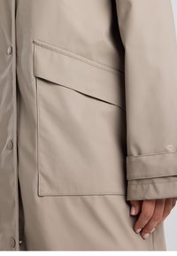 Manteau imperméable beige avec une grande poche à rabat, des boutons-pression et un détail de poignet, fabriqué dans un tissu lisse et léger.