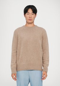 Kreminė megzta palaidinė su apvaliu kaklu ir gipiūrinėmis rankovėmis. Minkšta tekstūra, laisvas siluetas, dėvima su šviesiai mėlynomis dryžuotomis kelnėmis.