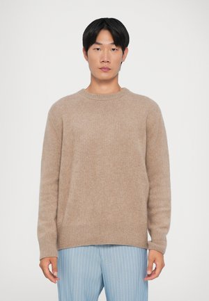 Beige pletený svetr s kulatým výstřihem a žebrovanými manžetami. Měkká textura, uvolněný střih, nošený s proužkovanými světle modrými kalhotami.