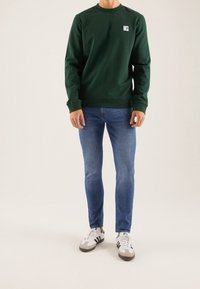 Felpa verde a girocollo con una piccola toppa con logo, indossata con jeans skinny blu e sneakers bianche con accenti grigi.