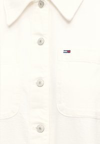 Chemise blanche boutonnée avec des boutons en métal gravés "Tommy Jeans" et un petit logo Tommy Hilfiger brodé sur la poche poitrine.