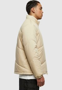 Beige pufferjakke med høy krage, quiltet design, elastiske mansjetter og løs passform. Tøyet ser glatt og isolert ut.