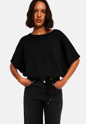 Kvinde med skulderlangt mørkt hår iført løs sort kortærmet top og sort højtbarmede jeans med hænderne i lommerne.
