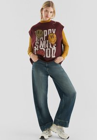 Vrouw in een bordeauxrood mouwloos vest met vrolijke gezichtjes en de tekst "Happy Smile Good", mosterdgele geribbelde lange mouwen top, brede jeans en witte sneakers.