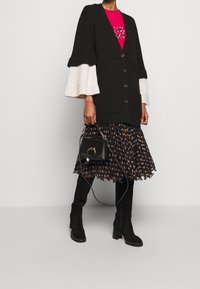 Sort ribbenstrikket cardigan med hvide klokkeærmer, sort blomsterkjole, sorte hælboots og en lille sort håndtaske med cirkel detalje.