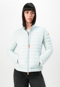 Vrouw met steil zwart haar die een lichtblauwe gewatteerde jas met oranje ritsen en zwarte broek draagt, met haar handen in haar zakken, kijkend naar rechts.